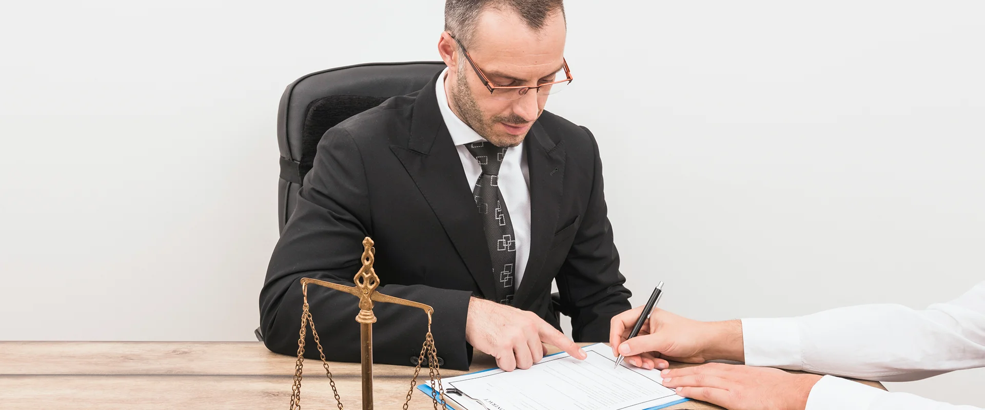 A lawyer in a suit pointing at a document for a client to sign at a desk with a scales of justice statue.