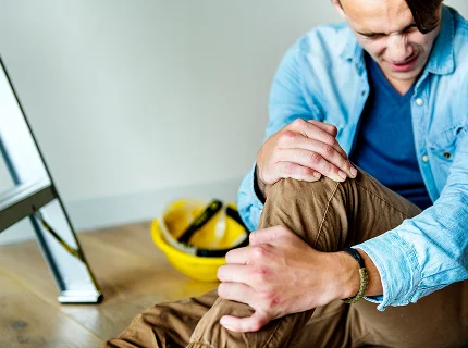 A man sitting on the floor grimacing in pain while holding his injured knee next to a yellow hard hat and ladder.